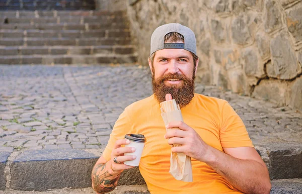 Cachorro-quente em movimento. Hipster descansando e tendo fast food cachorro quente durante a viagem de verão. Homem barbudo a comer sanduíche de cachorro quente. Caucasiano cara turista gostando de cachorro quente cozinha de rua — Fotografia de Stock