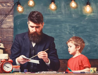 Sanat dersi kavramı. Sakallı öğretmen, küçük oğlu yakın, sınıfta, arka planda tebeşir tahtası takılıyor. Meşgul yüz boyama, çizim çocuk ve öğretmen