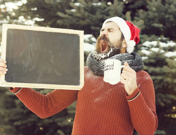 Bonito bocejo homem em chapéu de santa claus, hipster barbudo com barba e bigode coberto com geada branca, detém placa preta em branco e xícara de café dia de inverno ao ar livre em fundo natural, cópia spac — Fotografia de Stock