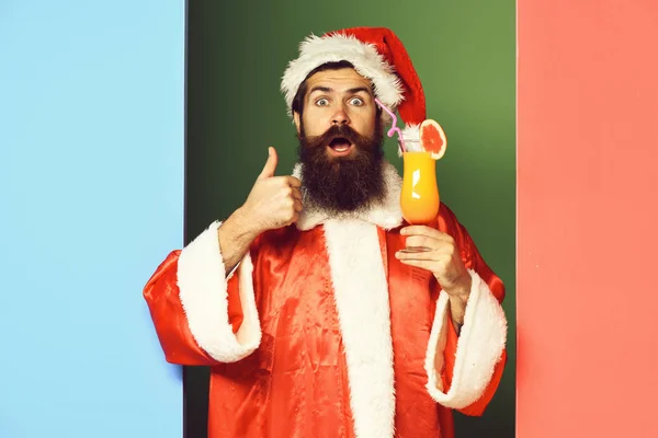 Schöner bärtiger Weihnachtsmann mit langem Bart im überraschten Gesicht, der ein Glas alkoholfreien Cocktails in Weihnachts- oder Weihnachtspullover und Neujahrsmütze hält und auf buntem Studiohintergrund cool wirkt — Stockfoto