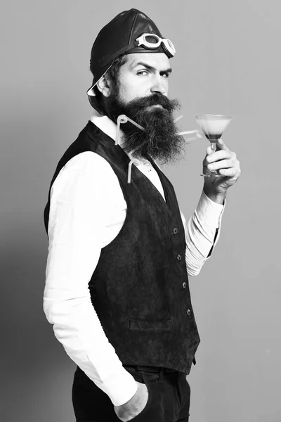 Handsome bearded pilot or aviator man with long beard and mustache on serious face holding glass of alcoholic beverage in vintage suede leather waistcoat with hat and glasses on red studio background — Stock Photo, Image
