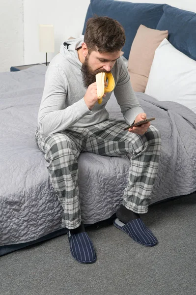 Brutal sleepy man has breakfast in bedroom. mature male with beard in pajama on bed. sleep and awake. healthy lifestyle. bearded man hipster eat banana in morning. Chatting with girlfriend — Stock Photo, Image