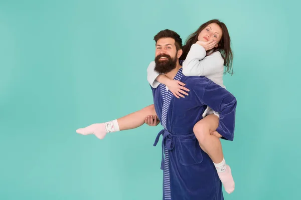 Close relationship. Handsome young man giving his girlfriend piggyback ride. Couple in bathrobes having fun turquoise background. Lets stay at home and have fun. They always have fun together