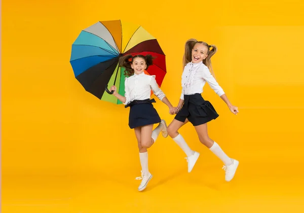 C'est un jour de pluie. Heureux petits enfants marchant à l'école sous la pluie sur fond jaune. Petites écolières souriantes avec parapluie de couleur arc-en-ciel. L'automne est la saison des pluies — Photo