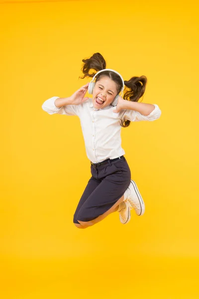 Música de la felicidad. Niño feliz saltando con felicidad sobre fondo amarillo. Feliz niña pequeña disfrutar escuchando música en los auriculares modernos. Frecuencia de felicidad. La felicidad ha vuelto a la escuela — Foto de Stock