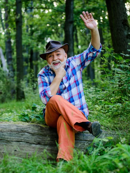 Olá. Homem maduro com barba de chapéu de cowboy. agricultor sentar-se em madeira. Feliz silvicultor. humano e natureza. caminhadas em madeira profunda. dono da floresta. piquenique de verão ou primavera. homem idoso agricultor relaxar na floresta — Fotografia de Stock