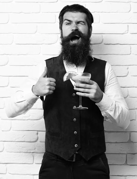 Handsome bearded man with long beard and mustache has stylish hair on funny face holding glass of alcoholic beverage in vintage suede leather waistcoat on white brick wall studio background — Stock Photo, Image