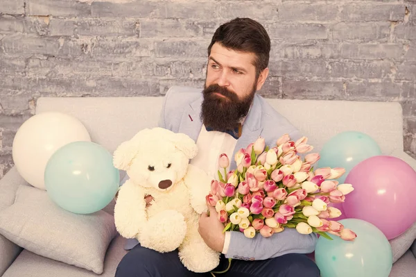 Romantic man with flowers and teddy bear sit on couch with air balloons waiting girlfriend. Romantic gift. Macho ready romantic date. Man wear tuxedo bow tie hold bouquet. She deserve all best