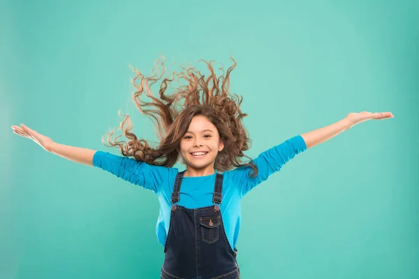 Erleben Sie ein Haar, um Ihre Energie auszugleichen. Entzückendes Mädchen mit lockigem Haar, das auf blauem Hintergrund wedelt. Kleines nettes Mädchen mit langen brünetten Haaren im lässigen Stil. Kleines Haar-Model mit Mode-Look — Stockfoto