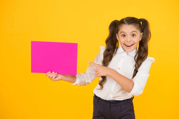 Voici votre produit. Bonne petite fille pointant du doigt le papier rose vierge pour la publicité de produits sur fond jaune. Produit de retour à l'école. Attirer l'attention sur le produit ou l'événement, espace de copie — Photo