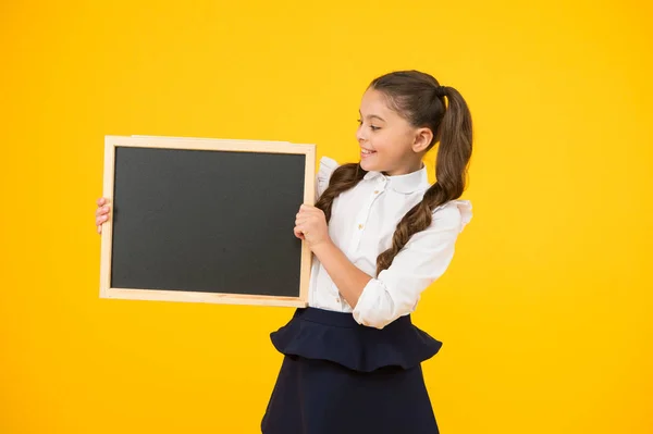 Menina pequena, grande sonho. Criança pequena segurando placa preta para propaganda escolar no fundo amarelo. Criança pequena com quadro-negro vazio para o seu texto. Pequena estudante segurando quadro negro, espaço de cópia — Fotografia de Stock