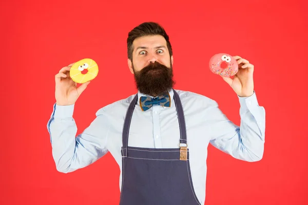 Кейтеринг хлібобулочних виробів для вас. Бородатий чоловік тримає кільцеві пончики в хлібопекарні. Професійний пекар з глазурованою хлібобулочною їжею на червоному тлі. Ідеальні пончики з нашої пекарні — стокове фото