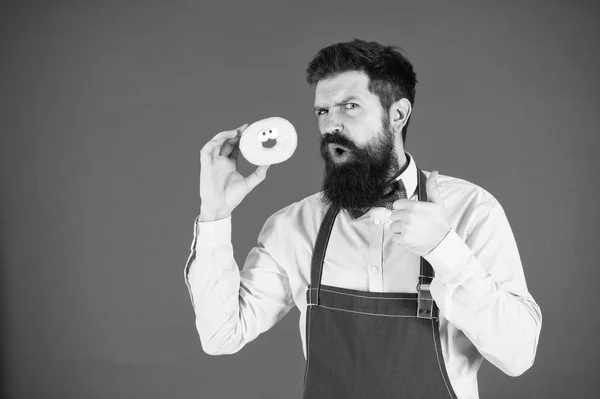 Hipster barbudo panadero mantenga rosquilla vidriada sobre fondo rojo. Concepto de cafetería y panadería. Un dulce donut de panadero. Hombre barbudo panadero en delantal celebrar lindo postre. Negocio privado de panadería. Inténtalo. — Foto de Stock