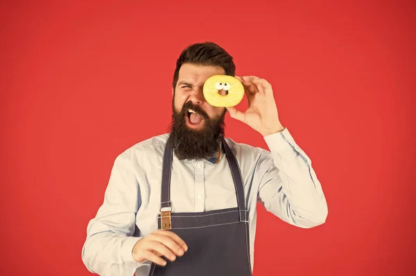 Oublie le régime. Manger le repas. Hipster barbu boulanger tenir beignet glacé sur fond rouge. Café et concept de boulangerie. Doux beignet du boulanger. Homme barbu boulanger dans tablier de cuisine tenir dessert mignon — Photo