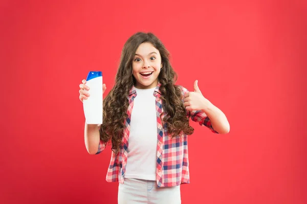 The best care ever. Little girl showing thumbs up for hair care product on red background. Happy small child liking her shampoo or gel for personal care. Skin and body care for kids