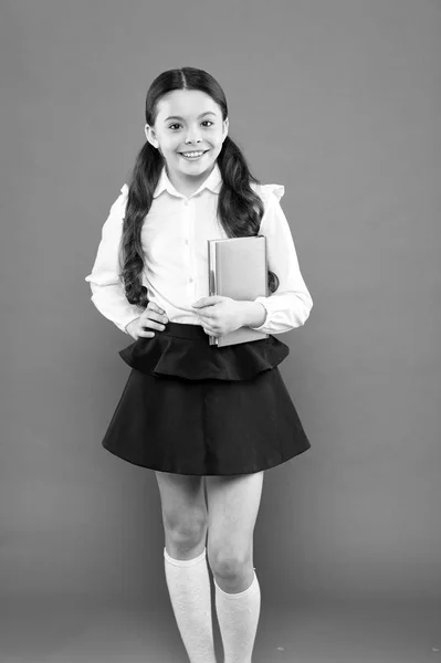 De volta à escola. Adorável menina desfrutando de seu tempo de escola em fundo laranja. Uma colegial feliz a ter aulas na escola primária. Bonito criança da escola sorrindo com o livro na mão — Fotografia de Stock