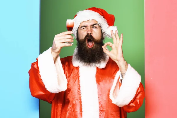 Schöner bärtiger Weihnachtsmann mit langem Bart auf lustigem Gesicht mit einem Glas alkoholischen Schnaps in rotem Weihnachtsmantel oder Weihnachtsmütze und Neujahrsmütze auf buntem Studiohintergrund — Stockfoto