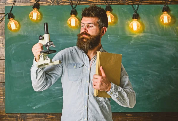 Profesor Hipster con microscopio, pizarra grande. Hombre con barba y bigote en cara estricta en clase. Concepto de investigación científica. Científico sostiene libro y microscopio, pizarra en el fondo — Foto de Stock