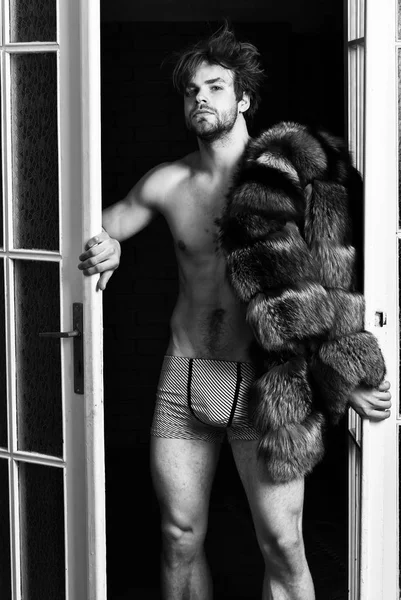 Luxury lifestyle and wellbeing. Luxury status symbol. Sexy macho tousled hair coming out bedroom door. Richness and luxury concept. Bachelor rich lover. Guy attractive posing fur coat on naked body