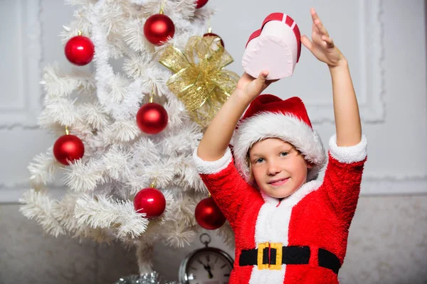 Świąteczny strój Świętego Mikołaja dla dziecka. Świąteczna impreza z stroju Świętego Mikołaja. Pomysły na choinkę dla dzieci. Chłopiec dziecko ubrany jak Mikołaj z czerwonym kapeluszem trzymać prezent pudełko w pobliżu choinki — Zdjęcie stockowe