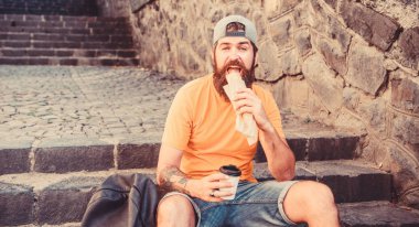 Adam sakallı hızlı atıştırmalık ve içecek kağıt fincan zevk. Sokak Yemekleri çok güzel. Kentsel yaşam tarzı beslenme. Merdivenlerde oturup Carefree hipster önemsiz yiyecek yemek. Aç adam atıştırmalığı. Önemsiz yiyecekler. Adam yemek sıcak köpek