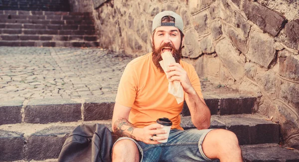 Adam sakallı hızlı atıştırmalık ve içecek kağıt fincan zevk. Sokak Yemekleri çok güzel. Kentsel yaşam tarzı beslenme. Merdivenlerde oturup Carefree hipster önemsiz yiyecek yemek. Aç adam atıştırmalığı. Önemsiz yiyecekler. Adam yemek sıcak köpek