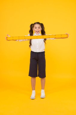 Ne uzun bir hükümdar. Sarı arka plan üzerinde sert ahşap cetvel tutan küçük öğrenci şaşırttı. Küçük çocuk metrik cetvel ile ölçüm alıyor. Cetvelim bir metre uzunluğunda.
