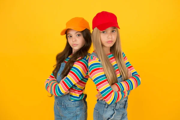 Moda che corrisponde al loro stile consapevole. Piccole ragazze in stile tenendo le braccia incrociate su sfondo giallo. Bambini che indossano cappelli da baseball in stile streetwear casual. Stile hip hop o hipster — Foto Stock