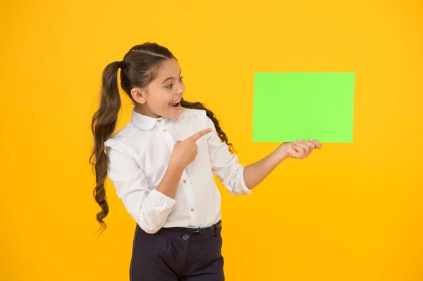 Gardez un œil sur l'idée. Fille heureuse pointant du doigt une certaine idée sur fond jaune. Petit enfant mignon souriant d'idée de génie. Petit enfant tenant du papier vide pour votre idée, copiez l'espace — Photo