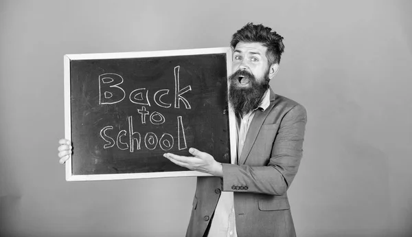 Man bearded in formal wear holds blackboard with text back to school. Clothing for teachers. School season advertisement. Man teacher classic clothing jacket and shirt stand over violet background