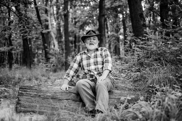 nature lover. senior man farmer relax in forest. mature man with beard in cowboy hat. farmer sit on wood. happy forester. human and nature. hiking in deep wood. forest owner. summer or spring picnic