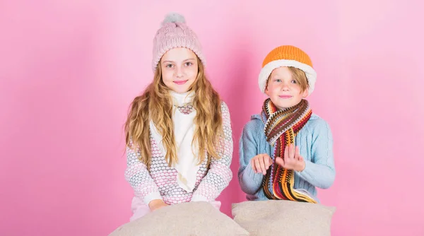 Bleiben Sie warm und bequem. wärmen Sie Ihre Winterbekleidung mit netten und gemütlichen Accessoires auf. Geschwister tragen winterwarme Hüte auf rosa Hintergrund. Kinder wärmen sich mit Kissen und Hüten auf — Stockfoto