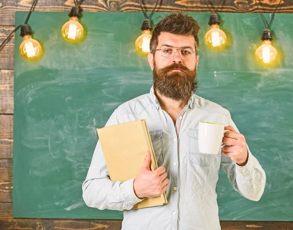 안경 을 입은 교사는 커피 나 차의 책과 잔을 들고 있습니다. 학교 휴식 개념입니다. 교실에서 차분한 얼굴에 수염을 가진 남자. 과학자는 커피의 책과 머그잔을 보유하고, 배경에 칠판, 복사 간격 — 스톡 사진