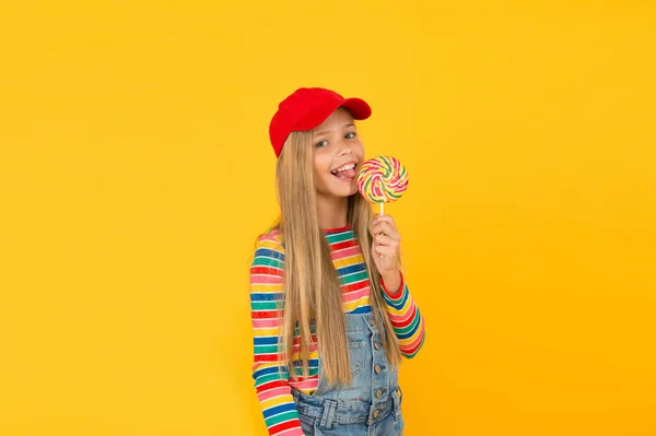 Il lecca-lecca la rende felice. Carina bambina che mangia caramelle vortice arcobaleno su sfondo giallo. Piccolo bambino leccare caramelle dure su bastone. Adorabile ragazzo felice che si gode il dolce di zucchero candito. Negozio di caramelle — Foto Stock