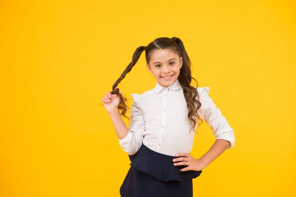Το διεστραμμένο της χτένισμα. Μικρό κορίτσι που τυλίγεις μακριά μαλλιά γύρω από το δάχτυλό της σε κίτρινο φόντο. Το μικρό παιδί με το χτένισμα της αλογοουρά στη σχολική στολή. Παιδί με νέο χτένισμα για πίσω στο σχολείο — Φωτογραφία Αρχείου
