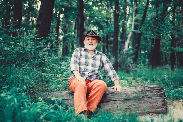 nature lover. senior man farmer relax in forest. mature man with beard in cowboy hat. farmer sit on wood. happy forester. human and nature. hiking in deep wood. forest owner. summer or spring picnic