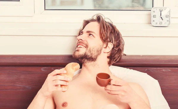 Bärtiger Mann im Bett, der morgens im Sonnenaufgangslicht Espresso trinkt. Der Mensch isst Croissant und trinkt Kaffee. Französisches Frühstückstereotyp. Brunch im Bett. — Stockfoto