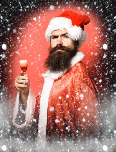 Guapo barbudo hombre de Santa Claus con barba larga en cara seria sosteniendo vaso de trago alcohólico en navidad o suéter de Navidad y sombrero de año nuevo en fondo de estudio rojo —  Fotos de Stock