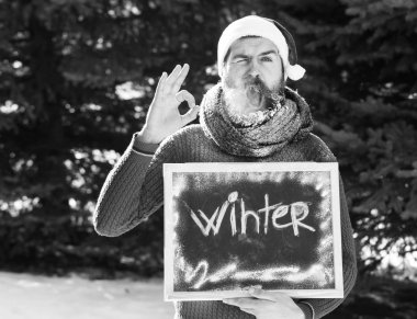 Santa claus şapka, sakal ve bıyık beyaz frost ile kaplı sakallı hippi neşeli adam kara tahta üzerinde kış kelime veya doğal adam güneşli günde kara tahta ok jestle verir