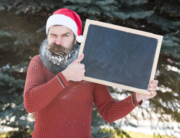 Knappe man van de ergens in Kerstman hoed, bebaarde hipster met baard en snor bedekt met white frost, houdt lege zwarte bord op winterdag buitenshuis op natuurlijke achtergrond, kopie vriendelij — Stockfoto