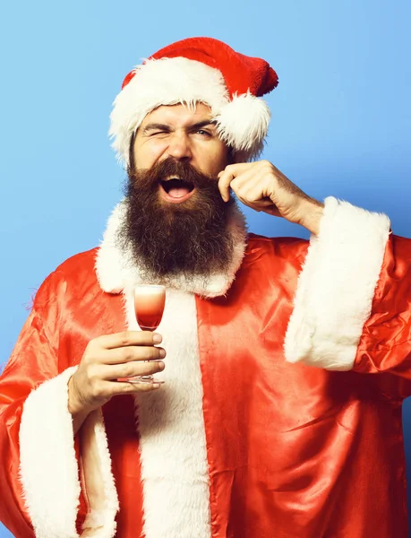 Knappe bebaarde Kerstman man met lange baard op grappig gezicht houden van glas alcoholische shot in Kerstmis of Xmas trui en Nieuwjaar hoed en het aanraken van snor op blauwe studio backgroun — Stockfoto