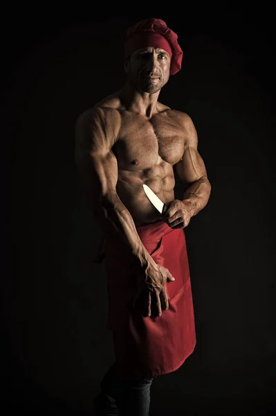 He is a mean cook. Professional master cook on black background. Man cook with muscular torso wearing chefs hat and apron. Chief cook with six pack abs holding kitchen knife