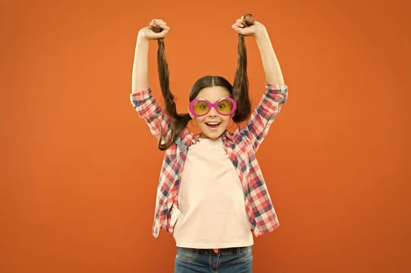 Porque es tu pelo. Adorable chica sosteniendo cola de caballo pelo sobre fondo naranja. Pequeña linda chica con el pelo largo morena usando gafas de lujo. Pequeño modelo de cabello con aspecto de belleza —  Fotos de Stock