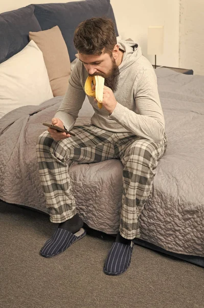 Hombre somnoliento brutal desayuna en el dormitorio. macho maduro con barba en pijama en la cama. Duerme y despierta. estilo de vida saludable. El hombre barbudo hipster come plátano por la mañana. Charlando con tu novia — Foto de Stock