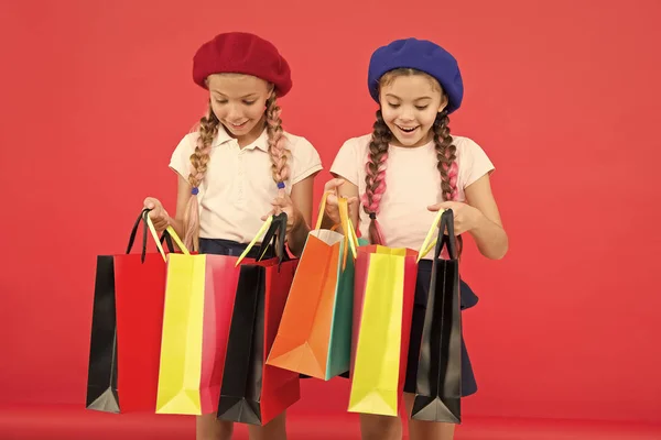 Niños lindas colegialas sostienen manojo bolsas de compras. Niños satisfechos comprando fondo rojo. Obsesionado con centros comerciales y de ropa. Concepto Shopaholic. Compras se convierten en diversión con los mejores amigos —  Fotos de Stock