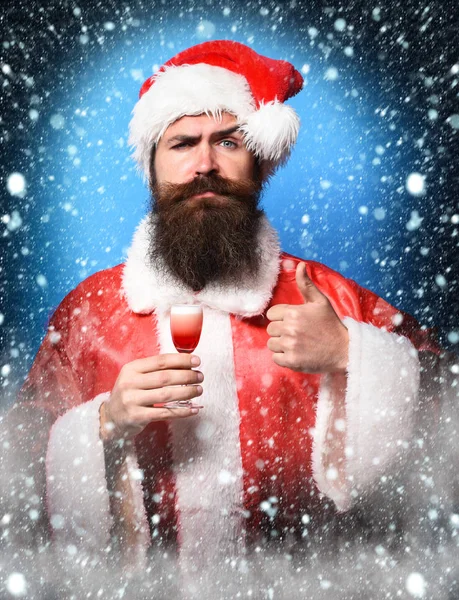 Handsome bearded santa claus man with long beard on serious face holding glass of alcoholic shot in red christmas or xmas sweater and new year hat showing cool on blue studio backgrou — Stockfoto