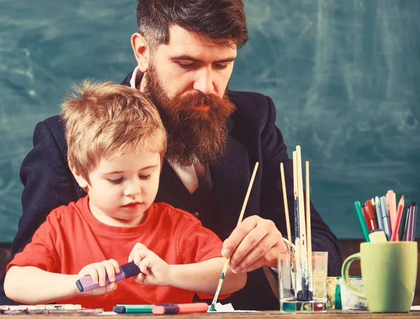 ひげを生やした教師、父と小さな息子が絵を描き、作成し、背景に黒板を描きながら教室で。才能と創造性の概念。忙しい顔の絵、ドローインの子供と教師 — ストック写真
