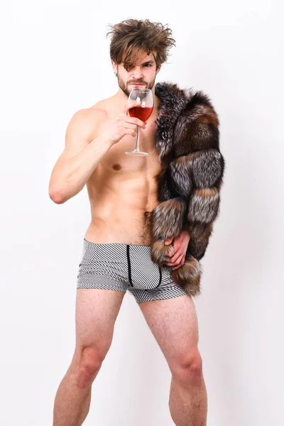 Riqueza y concepto de lujo. Chico atractivo posando abrigo de piel en cuerpo desnudo. Símbolo de lujo. Sexy macho soñoliento tousled beber vino o alcohol aislado en blanco. Estilo de vida y bienestar de lujo — Foto de Stock