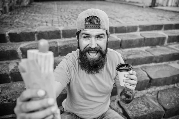 Lanche de bom humor. Um tipo a comer cachorros quentes. Homem barbudo comer salsicha saborosa e beber copo de papel. Conceito de comida de rua. Nutrição de estilo de vida urbano. Comida de plástico. Despreocupado hipster comer junk food enquanto sentar escadas — Fotografia de Stock