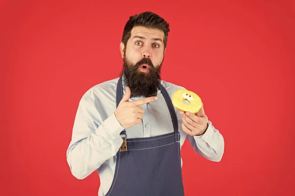 Doux beignet du boulanger. Homme barbu boulanger dans tablier de cuisine tenir dessert mignon. Façons de réduire la faim et l'appétit. Hipster barbu boulanger tenir beignet glacé sur fond rouge. Café et concept de boulangerie — Photo